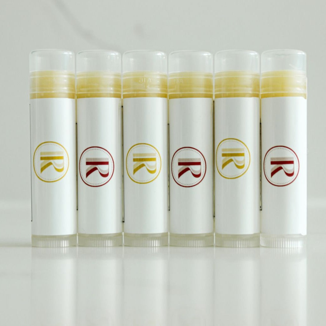 Elevate your lip care routine with our exquisite collection of clear lip balm tubes. Proudly displayed against a pristine white quartz countertop, these six standing tubes showcase a harmonious blend of clarity and sophistication. Each tube features a distinctive white label adorned with our logo, alternating between vibrant red and sunny yellow accents. The minimalist design exudes a sense of purity and quality. Explore our natural skincare line where simplicity meets elegance. Lip care, redefined.